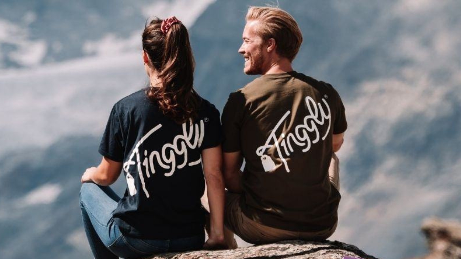 two individuals in a romantic relationship are sitting side by side on a hilltop, wearing t-shirts with the brand 